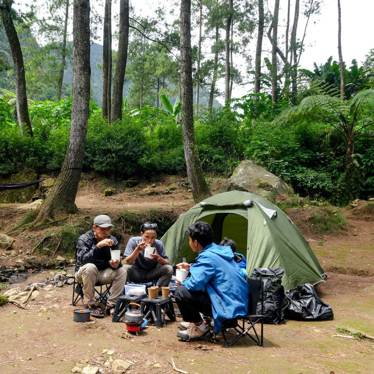 Three people camping in a forest with a green tent and chairs.