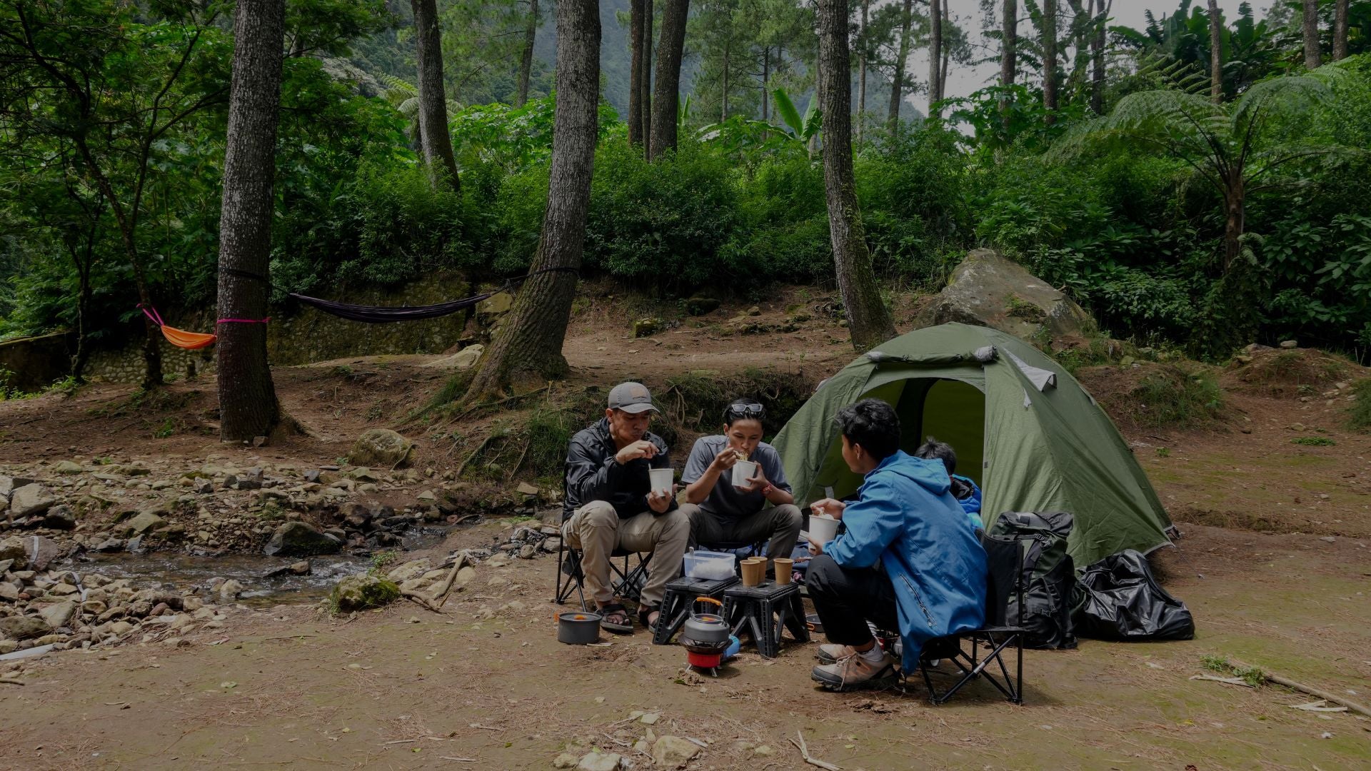 Three people camping in a forest with a green tent and chairs.